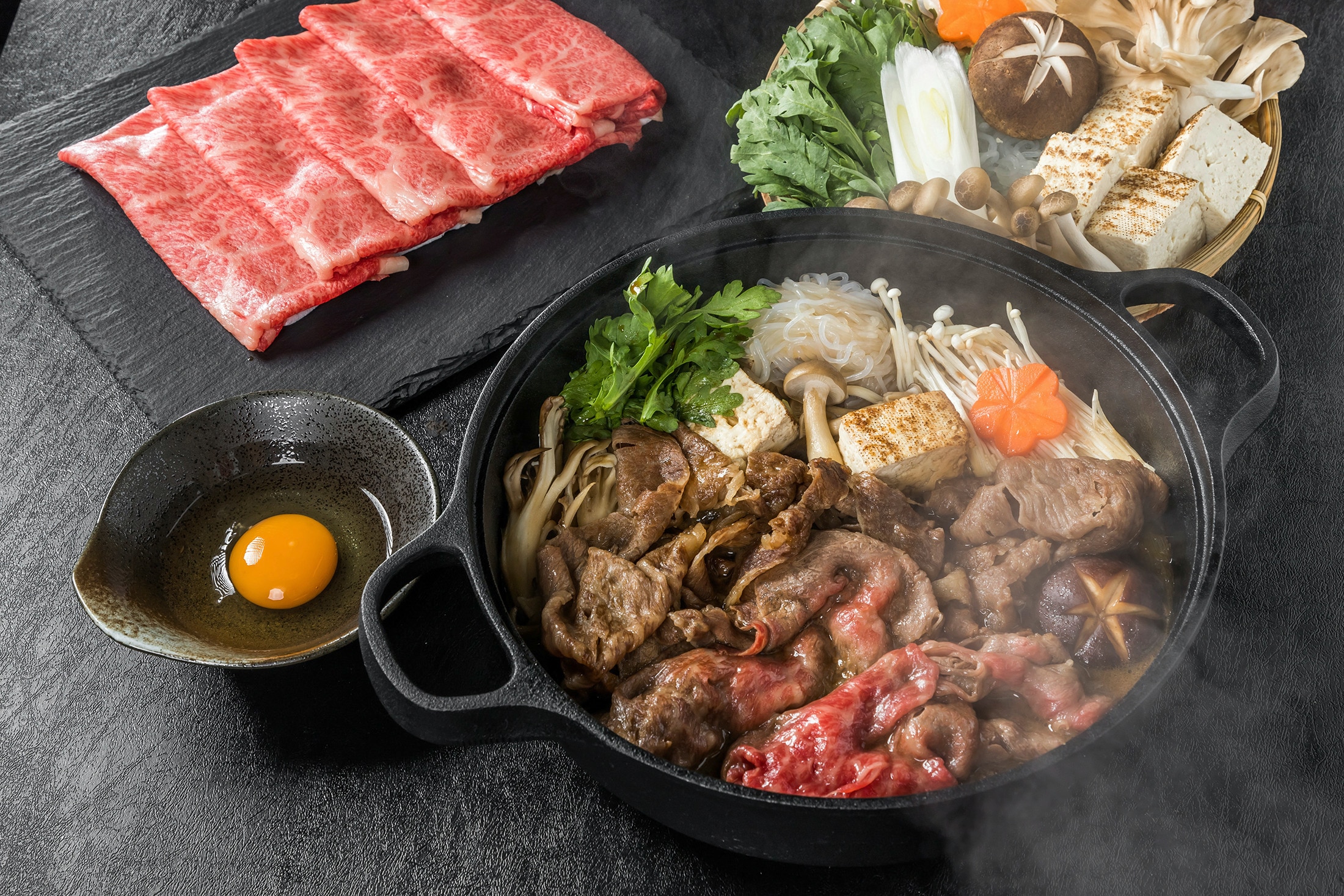 Sukiyaki in a cast-iron pan beside a saucer of raw egg and a platter of uncooked meat and vegetables