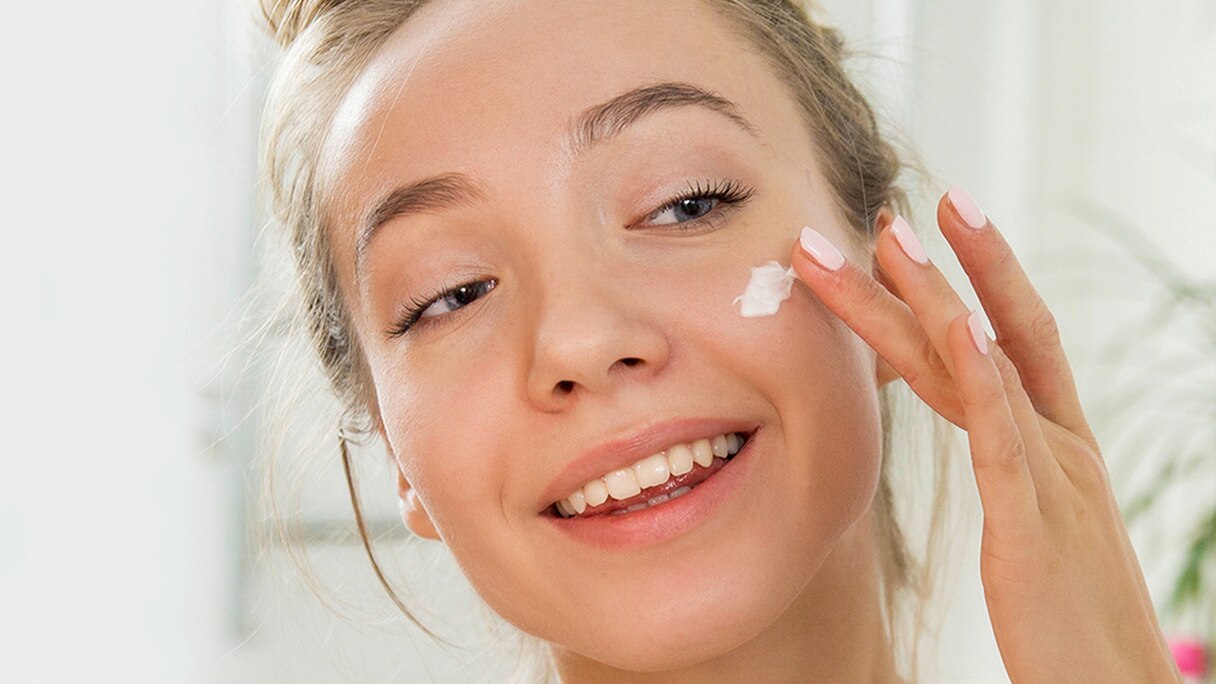 Smiling girl applying moisturiser to her cheek in a bathroom setting