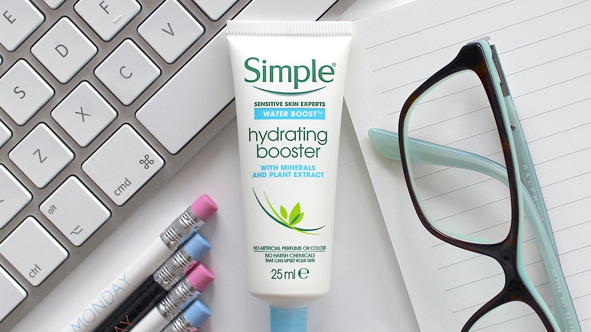 A tube of Simple Hydrating Booster on a white table with pencils, a keyboard and spectacles alongside it