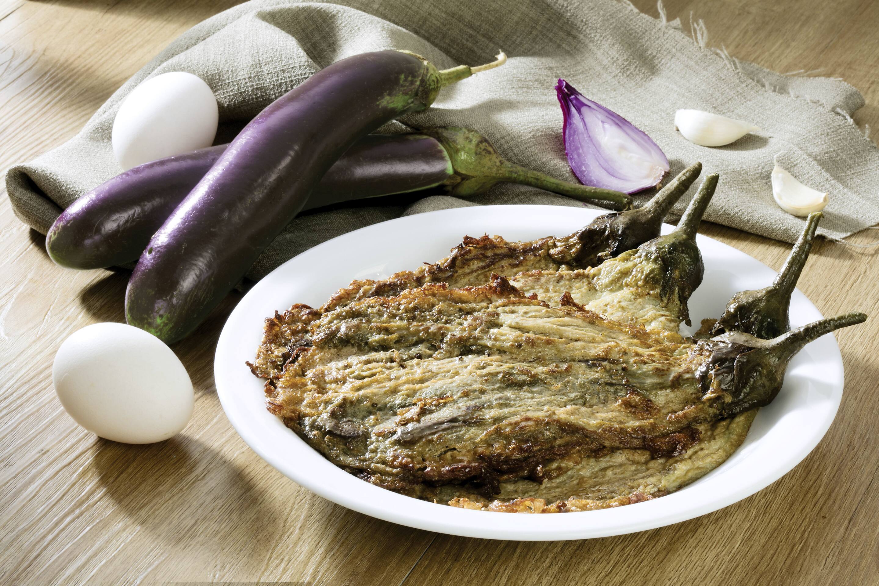 A plate of basic tortang talong, styled with eggs, raw eggplants, onion, and garlic cloves
