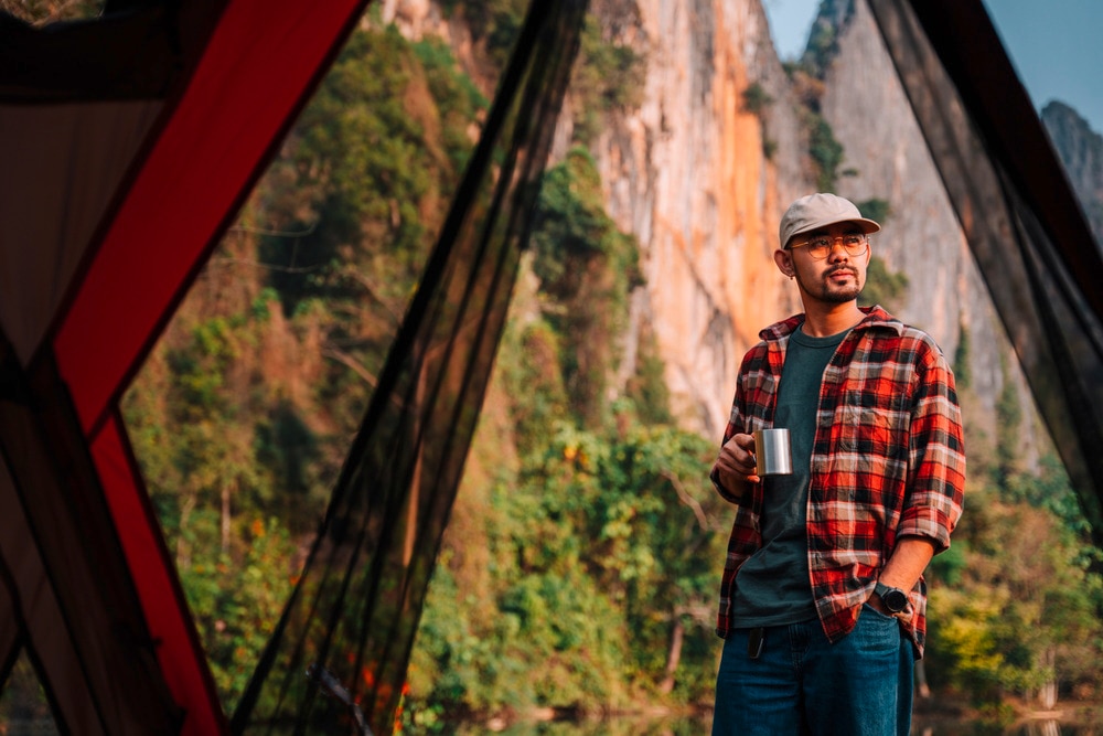 Man with a cap and flannel shirt looking at the view from a campground while holding a mug.
