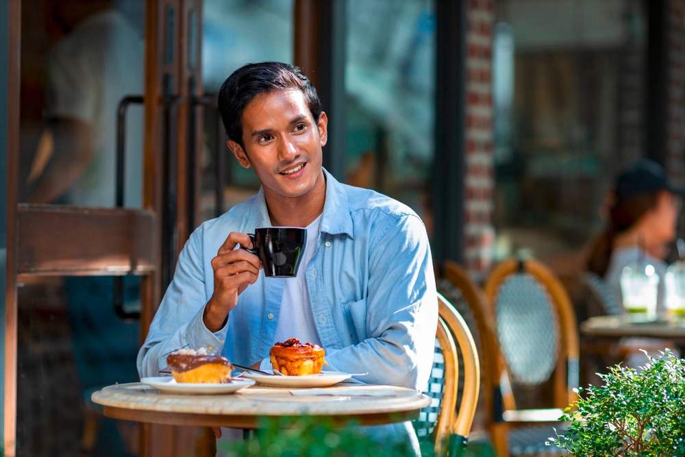 Man holding a black cup at a café with pastries on his table.