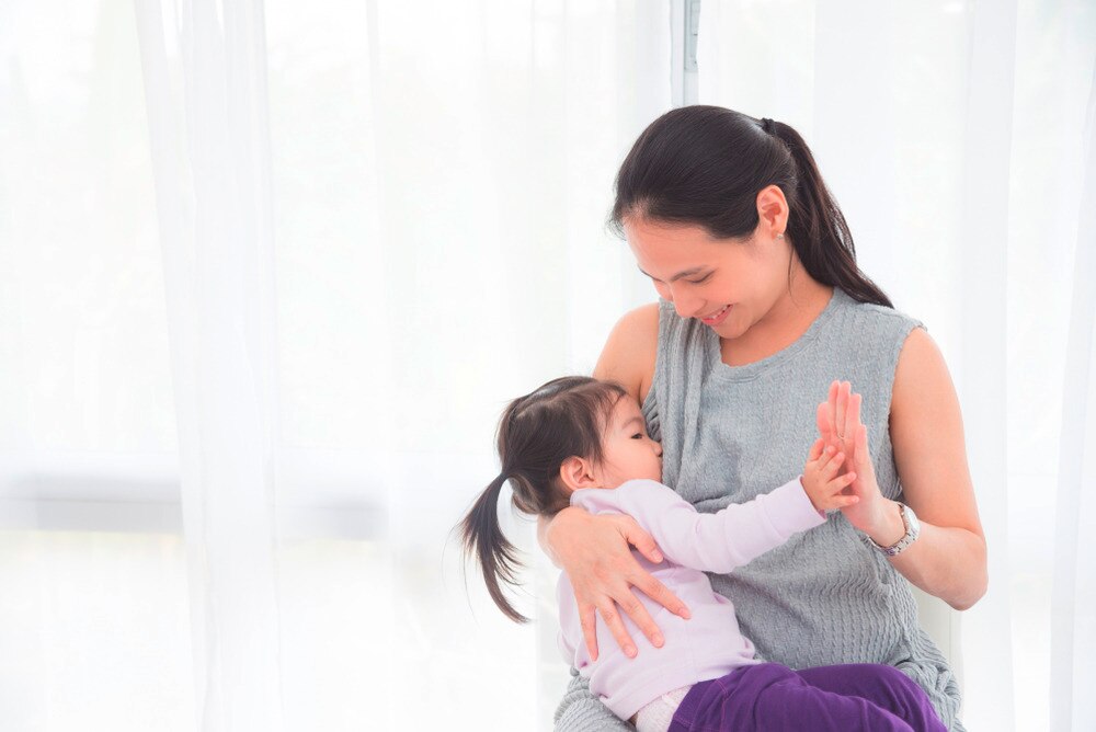 Asian mom in gray breastfeeding a female toddler.