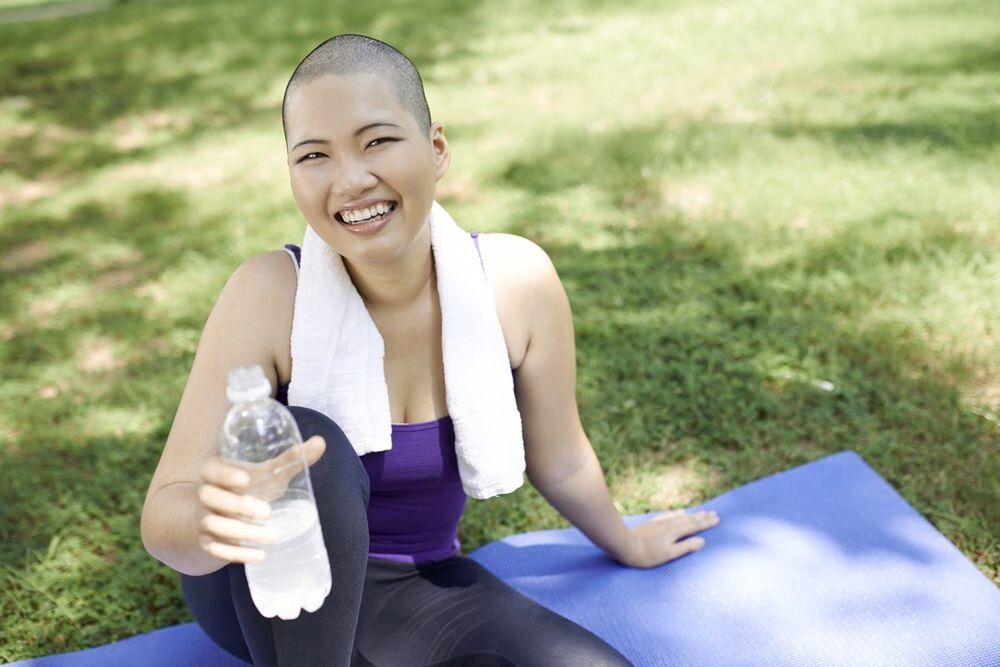 Asian girl smiling and holding a bottle of water on a yoga mat outdoors.