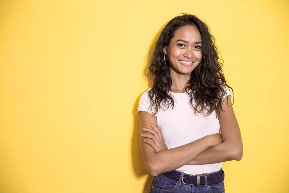 Pretty Asian girl with curly hair smiling and folding her arms against a yellow background.