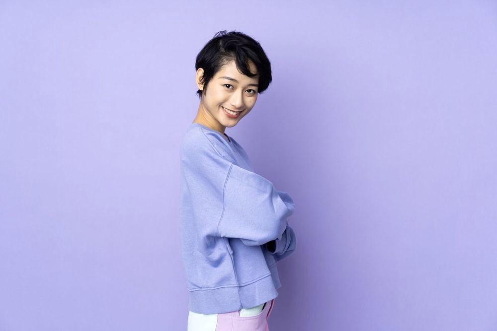 Asian woman smiling and folding her arms against a purple background