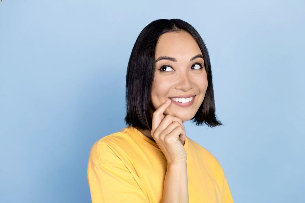 Closeup of an Asian woman smiling and putting her finger on her mouth against a blue background.