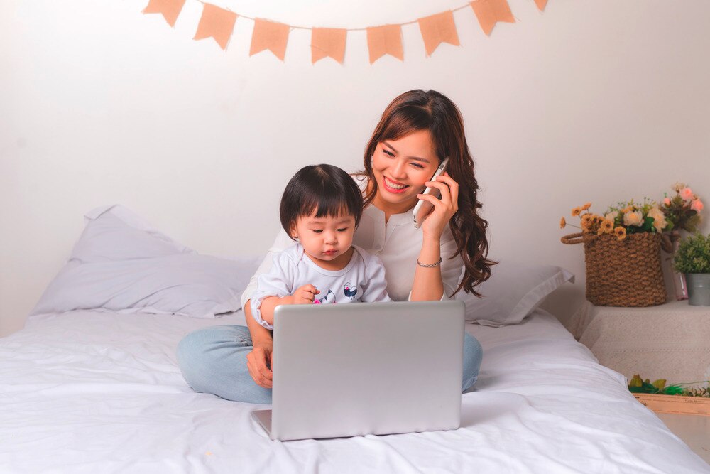 Asian mom working from a bed with her small child.