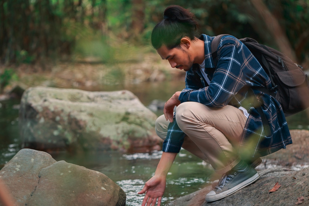 Asian hiker dipping his hand into a river in the forest.