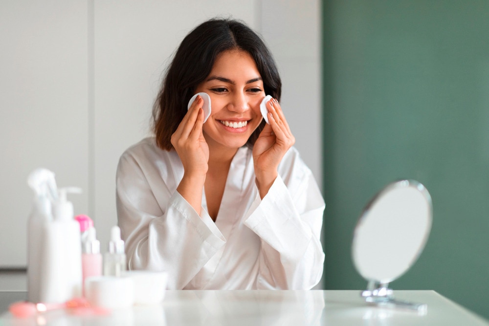 Woman rubbing her face with cotton pads while looking at a mirror. 