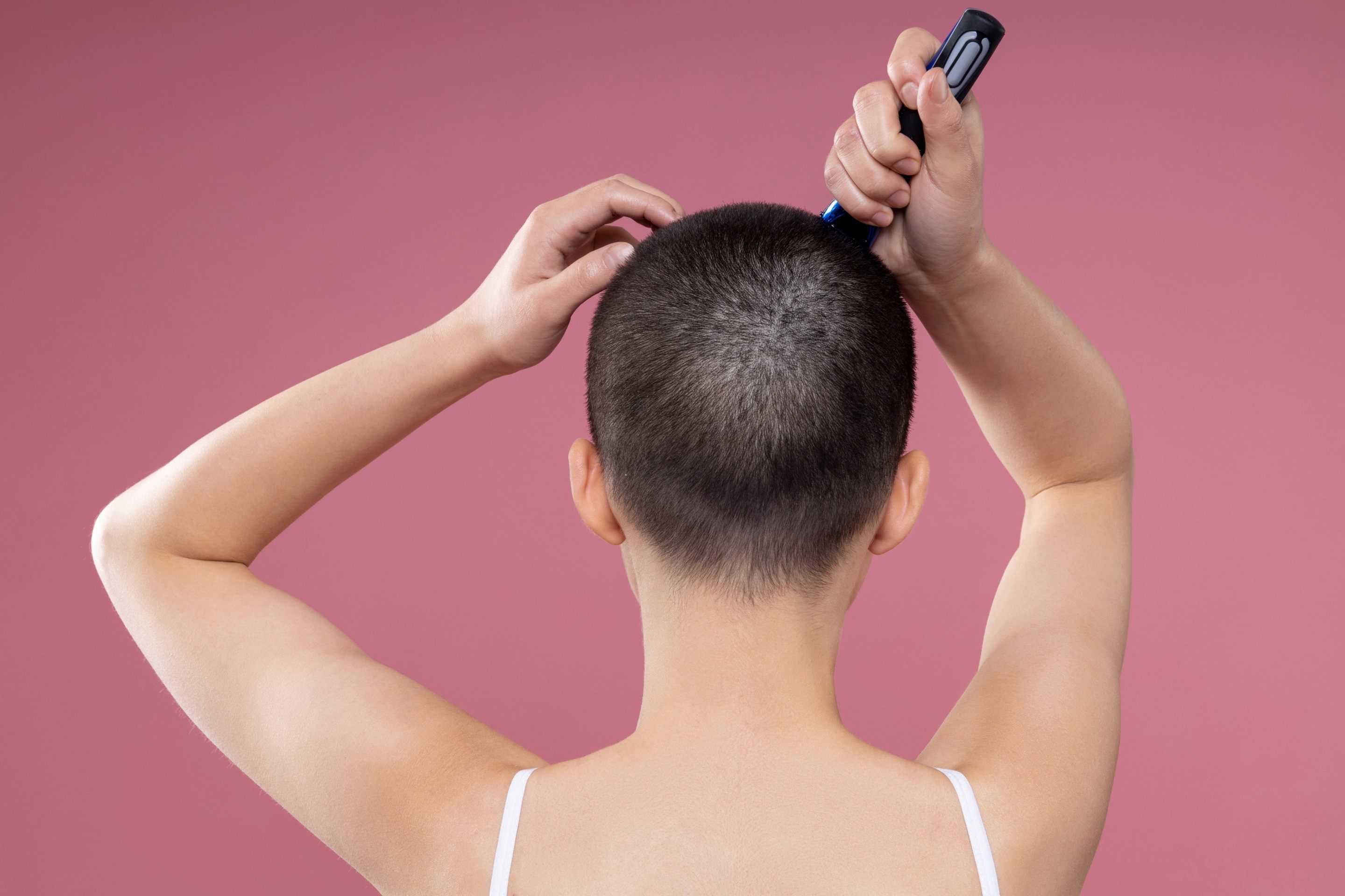 Rear view of woman shaving her head against pink background. 