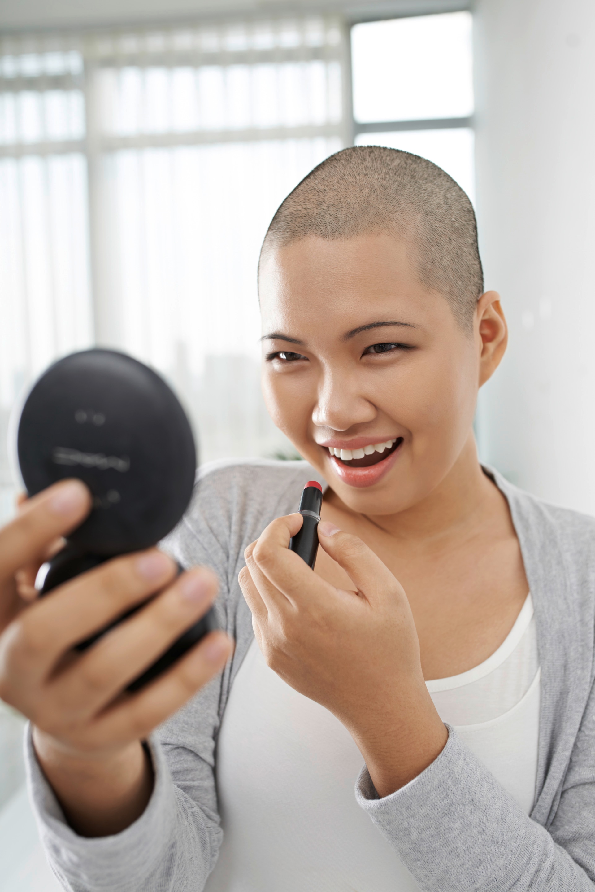 Asian woman with shaved head happily applying lipstick in front of a compact mirror. 