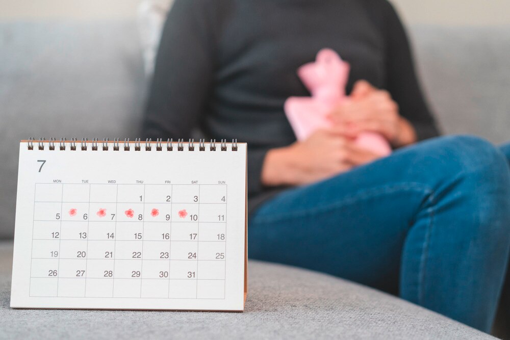 Woman soothing her stomach ache with a warm compress, a calendar marked with her period cycle on the table.