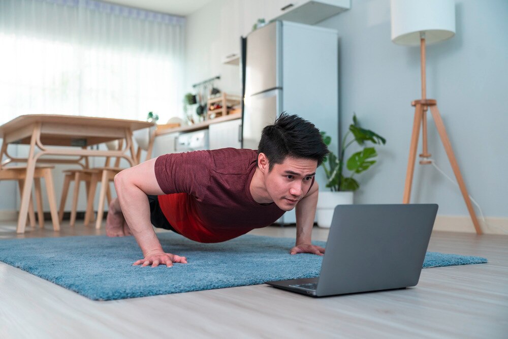 Young Asian male performs a push-up while watching on his laptop in his living room.