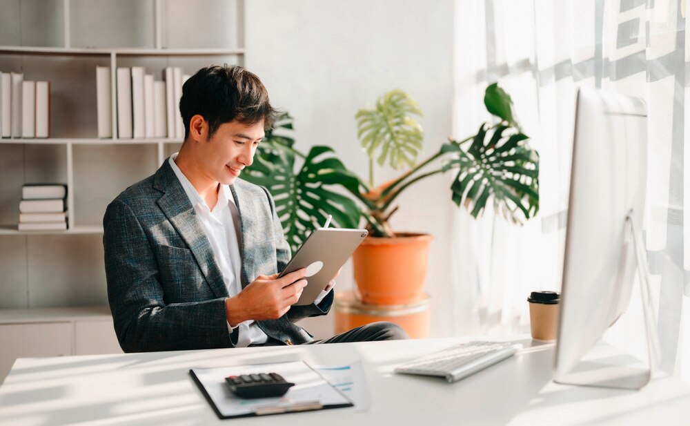 Young Asian male checks his tablet in front of the computer and a desk with his notes and calculator.
