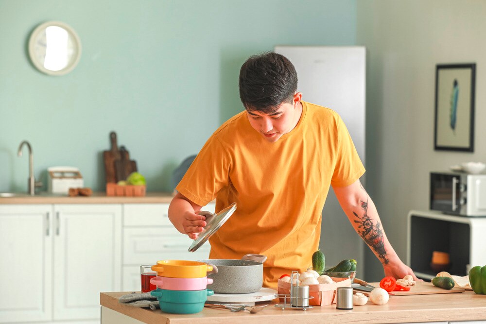 Young Asian male opens a pot's lid while ingredients are laid out on the kitchen counter.