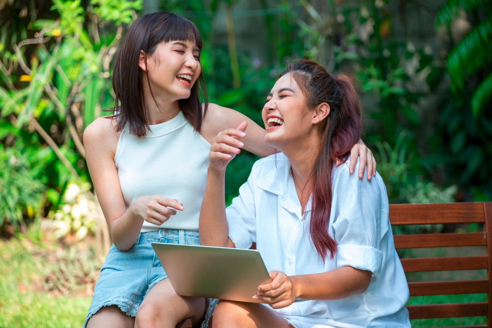 Two women laughing together while sitting on a bench with a laptop on her lap.