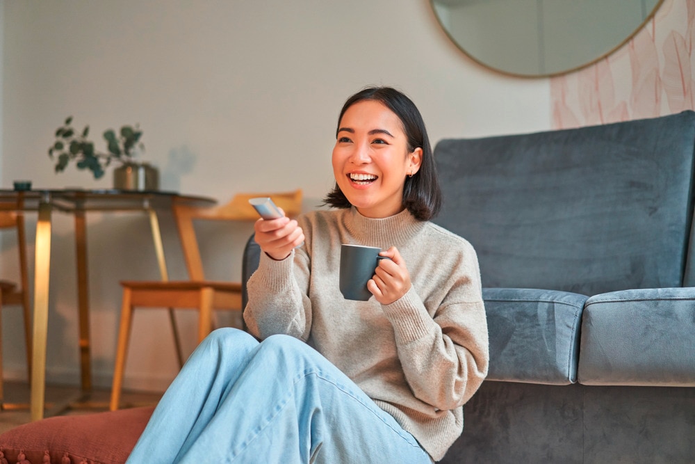 Asian woman laughing while holding a remote and watching TV.