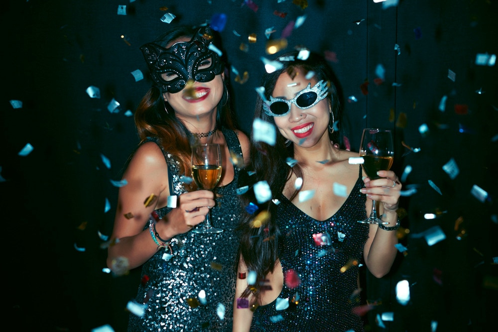 Two women in sparkly dress and masks holding glasses of water.