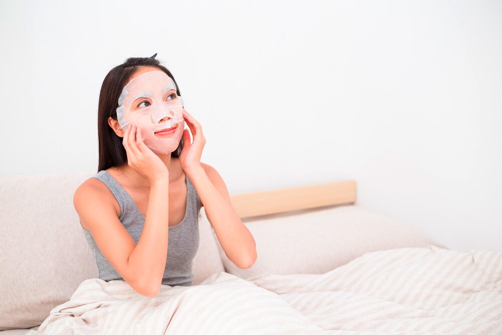 Woman wearing sheet mask on her face while sitting on a bed.