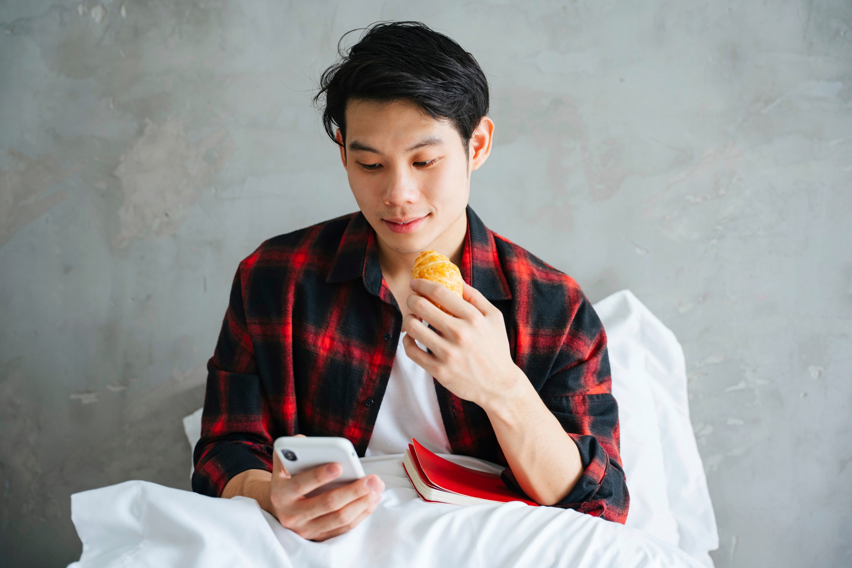 Asian man in red plaid eating bread in bed while holding cellphone