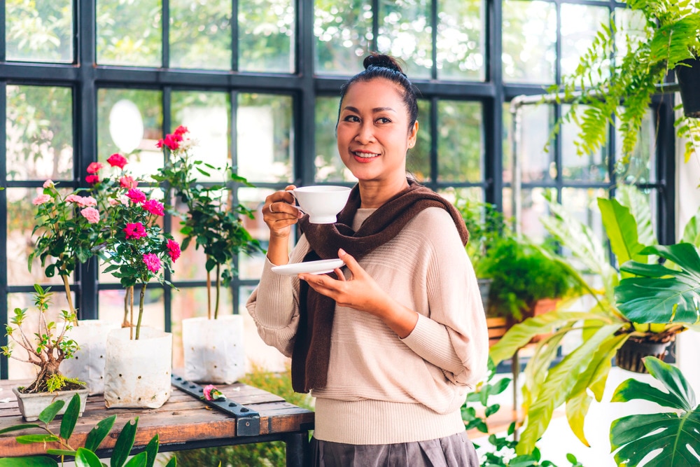 Woman drinking a cup of hot cocoa inside a plant-filled greenhouse.