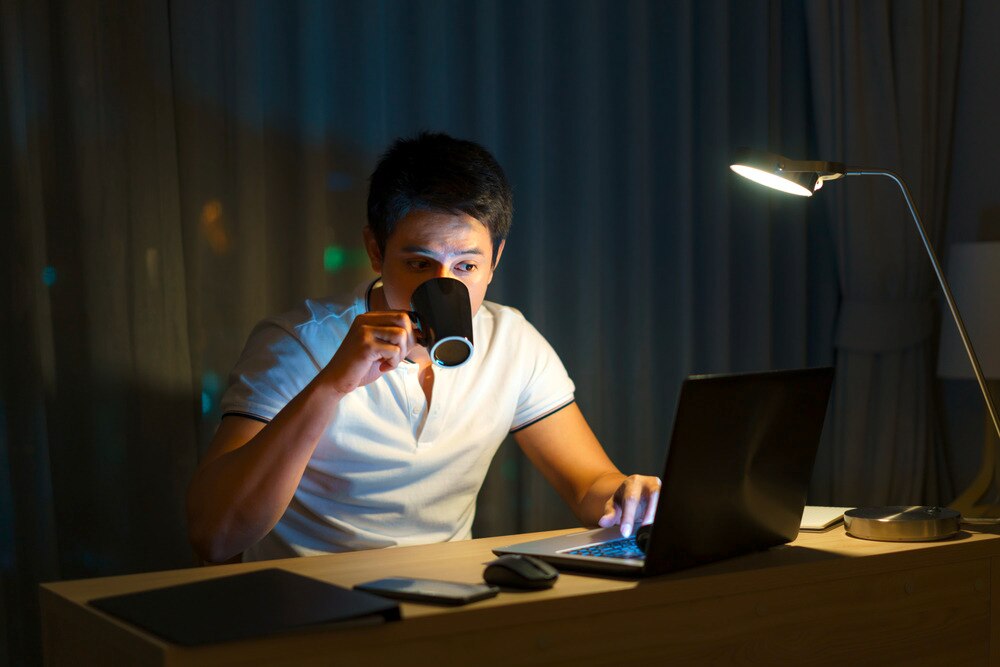 Man drinking from his cup while working on his laptop late at night.