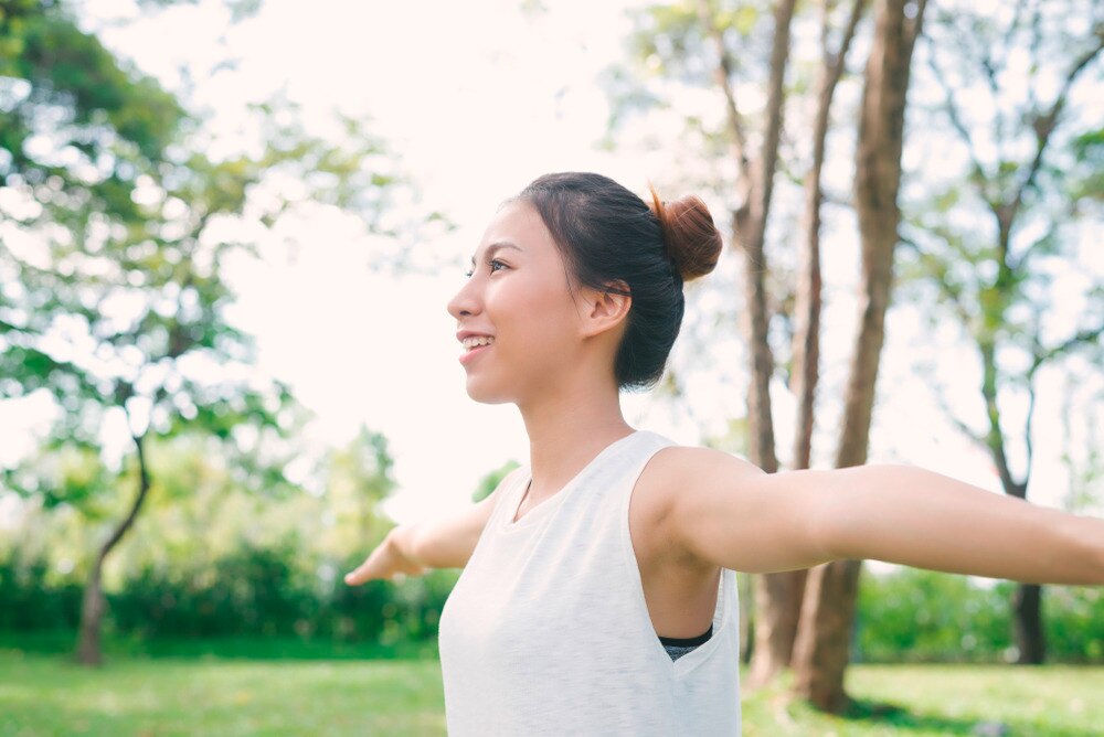 Asian woman in a white sleeveless shirt stretching before a workout outdoors.