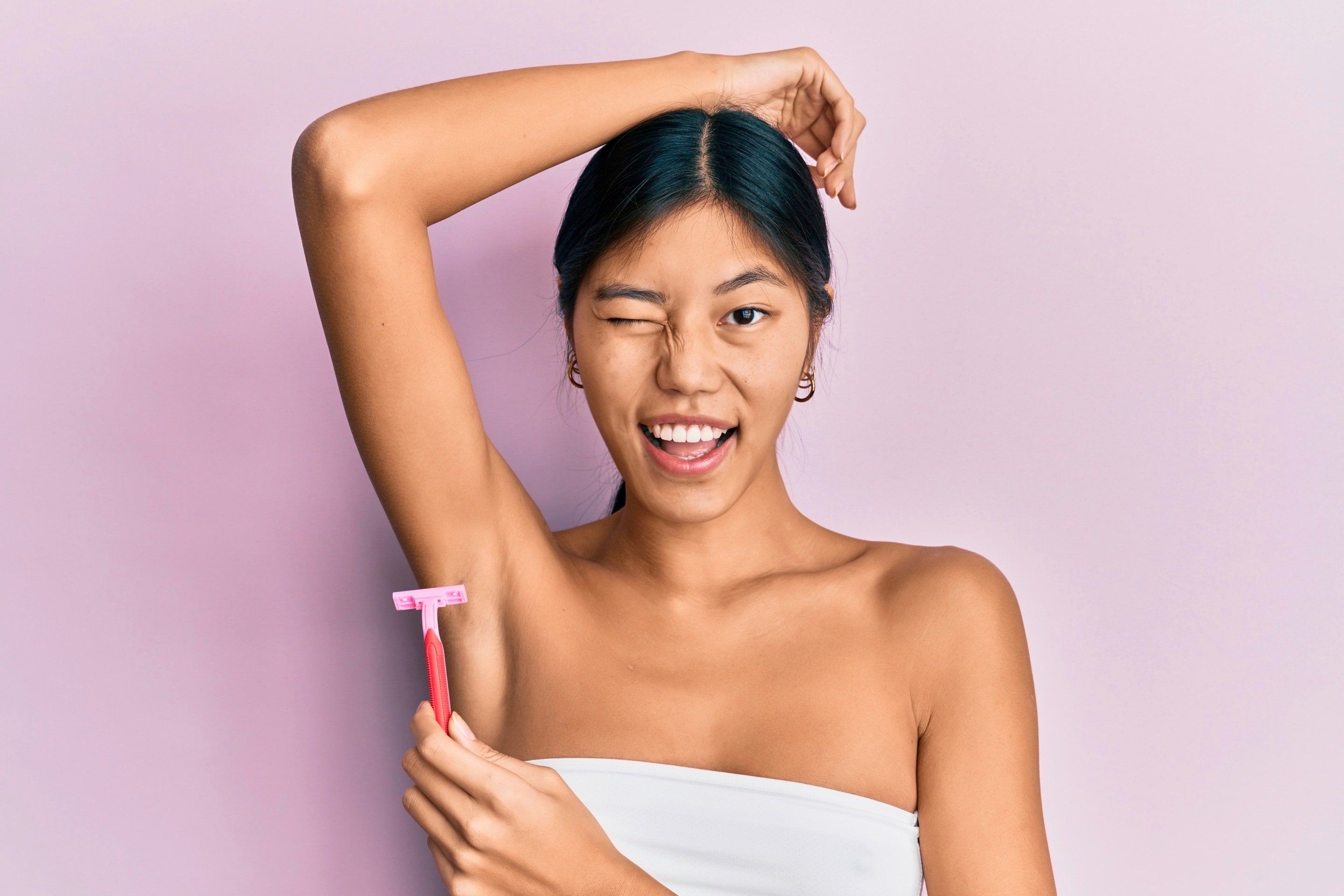 A tanned Asian woman with one arm raised, wearing a towel, and holding a pink razor