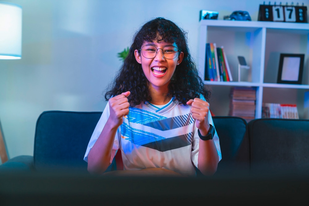 Woman with glasses and curly hair watching TV cheerfully at night.