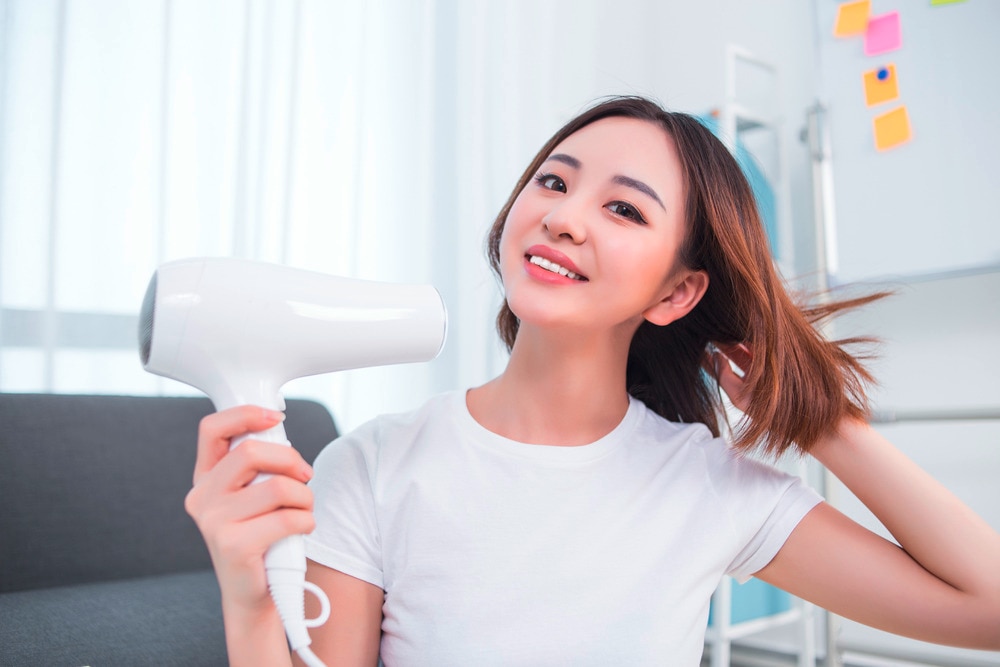 Smiling woman in white T-shirt drying her hair with a hair dryer.
