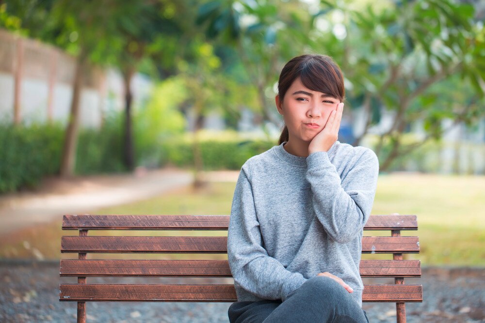Woman wearing gray sweater touching her cheek in pain while sitting on an park bench.
