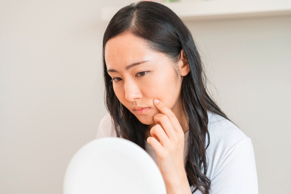 Woman touching her skin while looking at a mirror with a concerned expression.