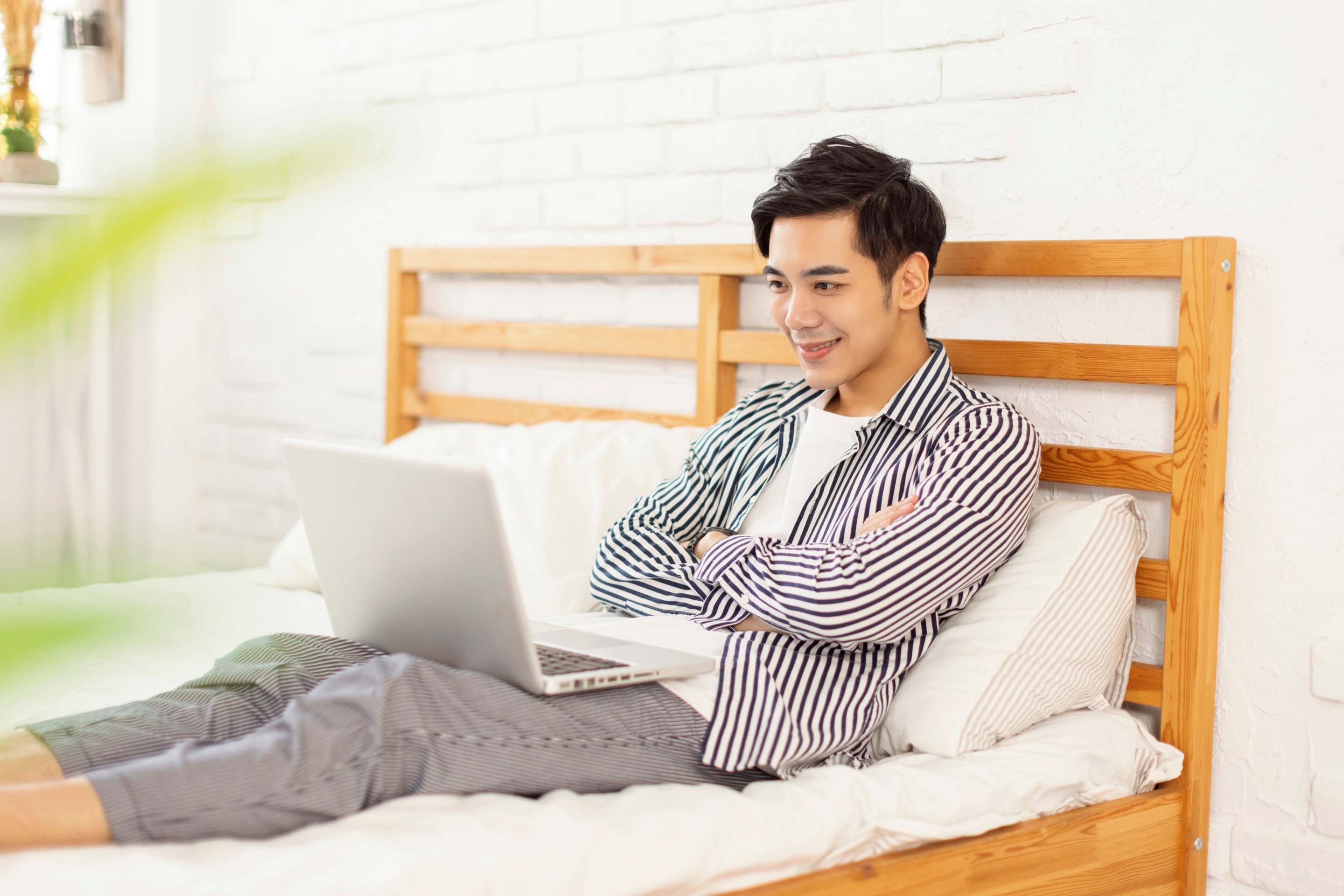 Asian man in jeans and striped shirts looking at laptop in bed