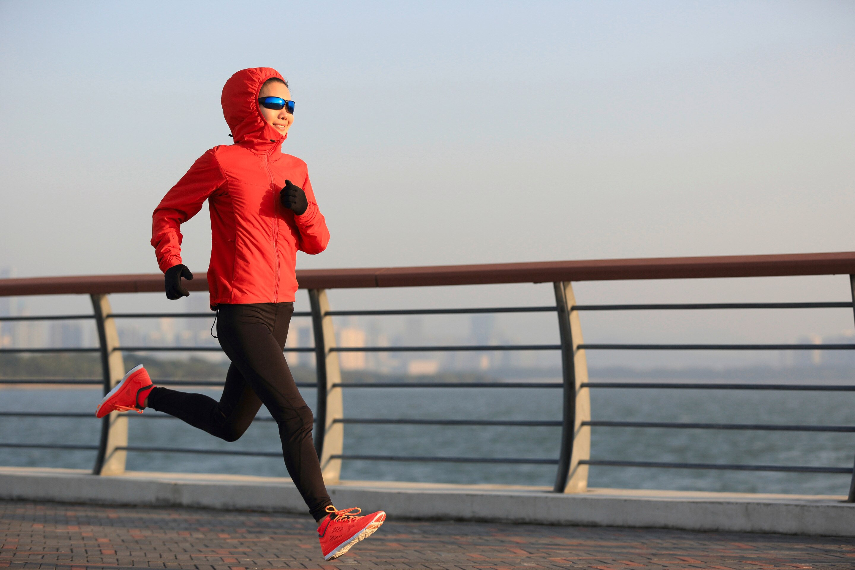 A woman wearing sunglasses, a red hoodie, and gloves jogging outside