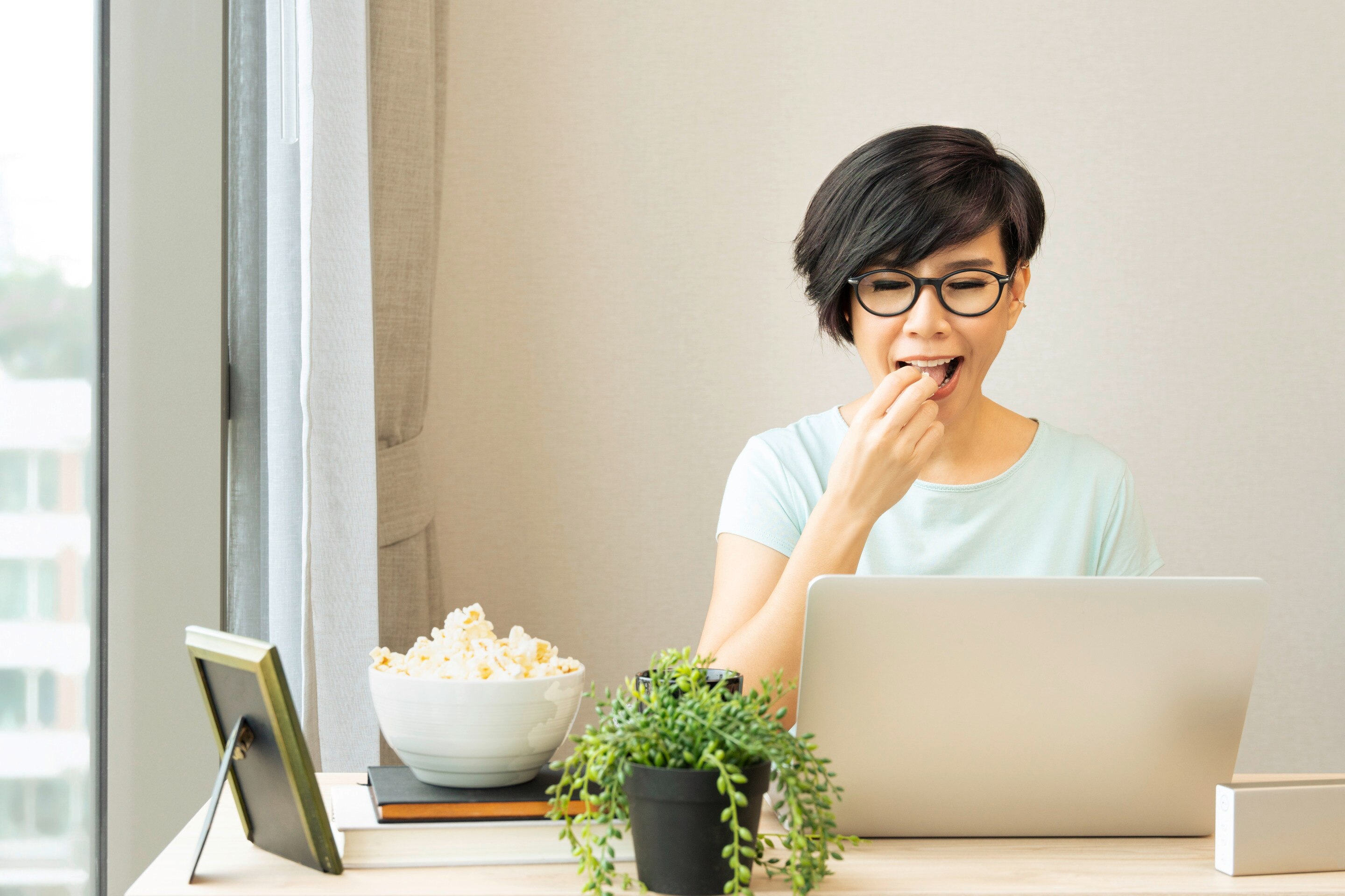 Asian woman with pixie cut and glasses having popcorn while working behind a laptop