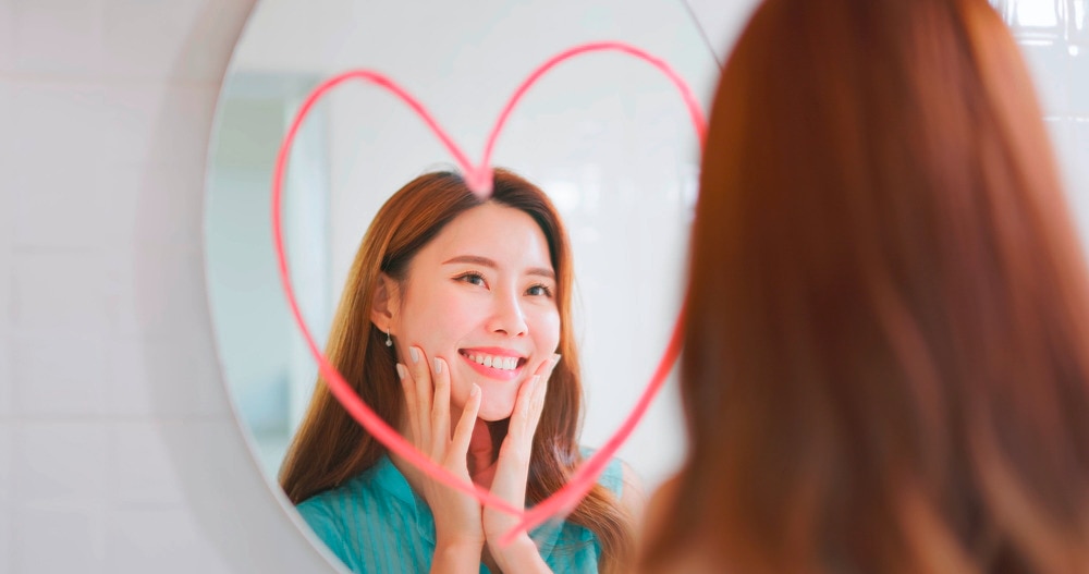 A woman smiling at herself in a mirror with a drawing of a heart on it.