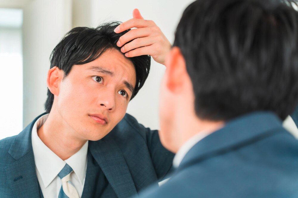 Asian man in suit looking at scalp in front of mirror.