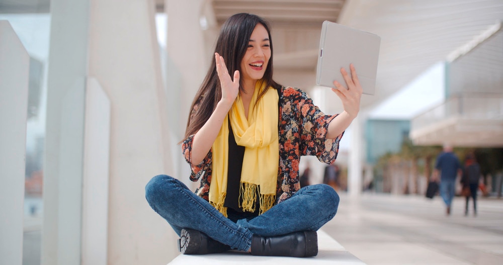 Asian woman wearing yellow scarf saying hello to a tablet.