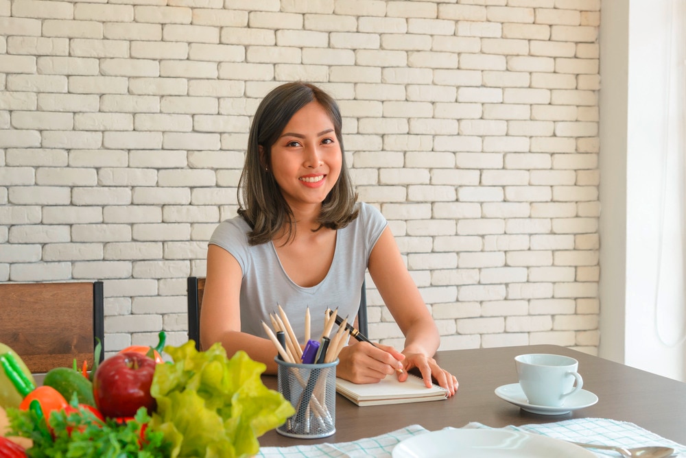 Asian woman writing on notebook in front of white brick wall.
