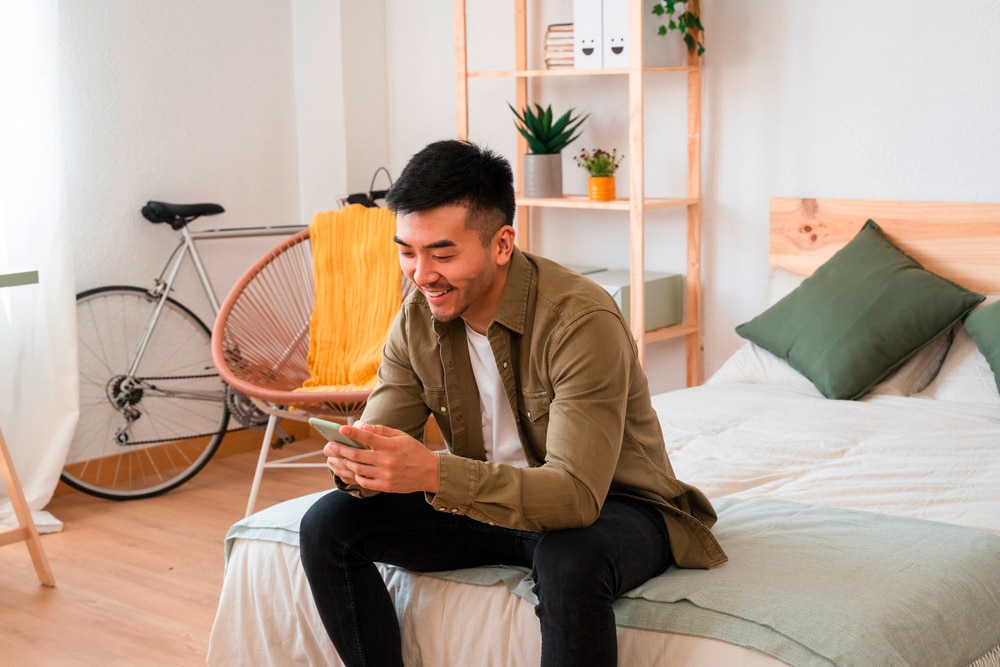 Man in smart casual outfits playing on his phone while sitting on the bed.