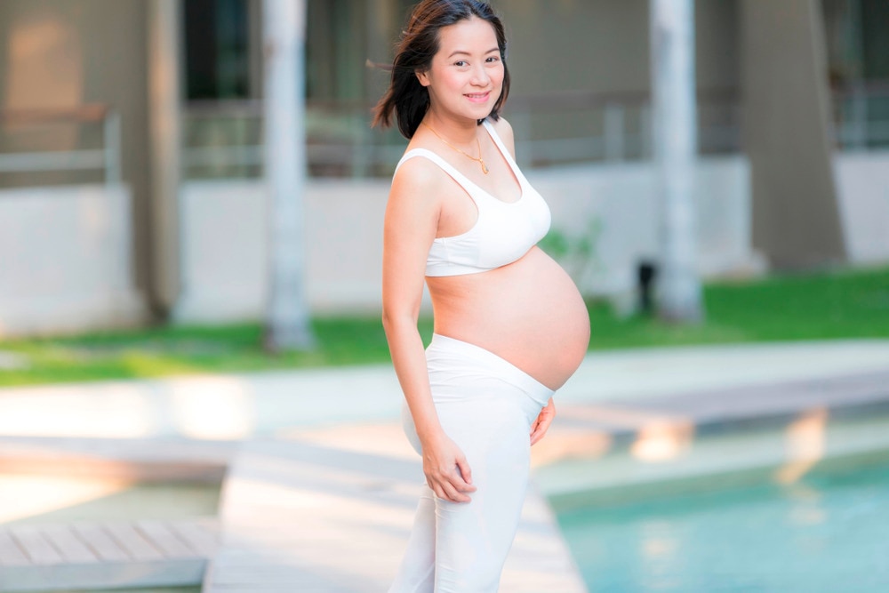 Pregnant woman in a white bra top and maxi skirt posing by the pool for her maternity shoot.
