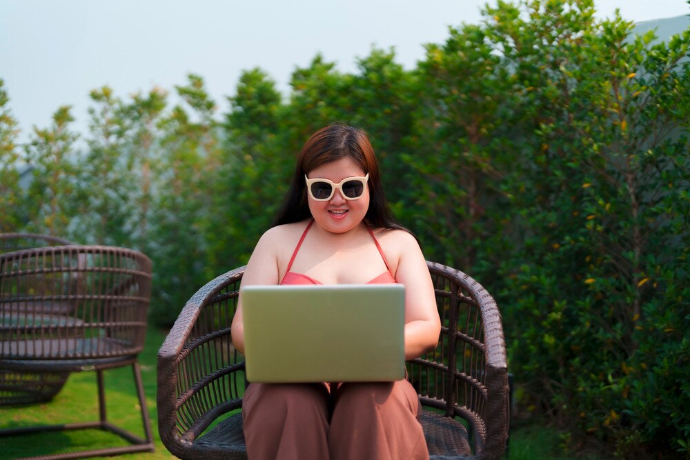 Happy, plus-size woman in sunglasses types on her computer outdoors.