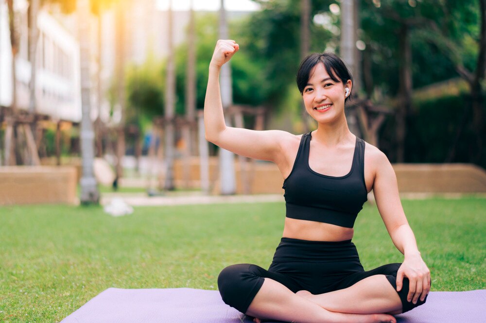 Woman sitting on a yoga mat outdoors, flexing her arm muscles.
