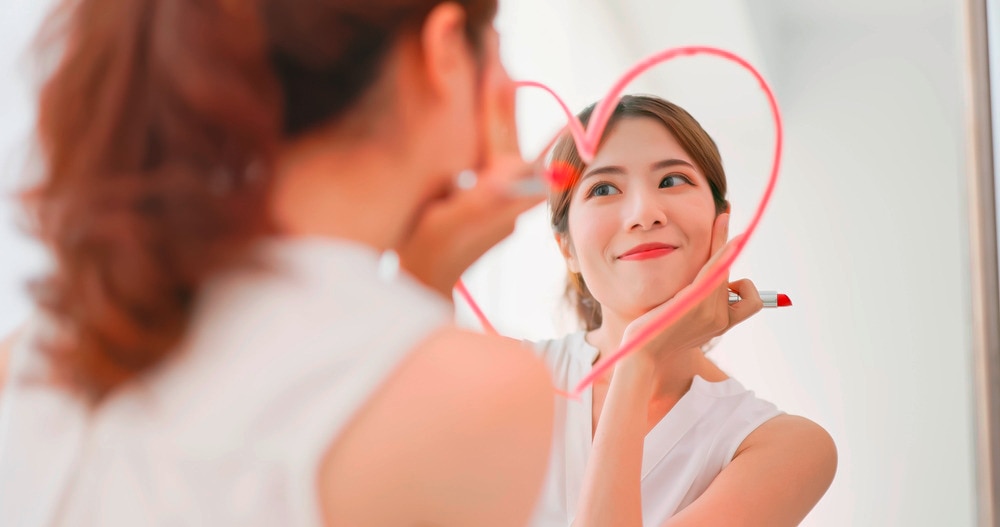 An Asian woman draws a heart on a mirror with lipstick.