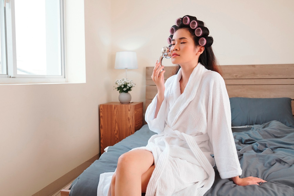 Woman in bathrobe wearing hair rollers while massaging her face.