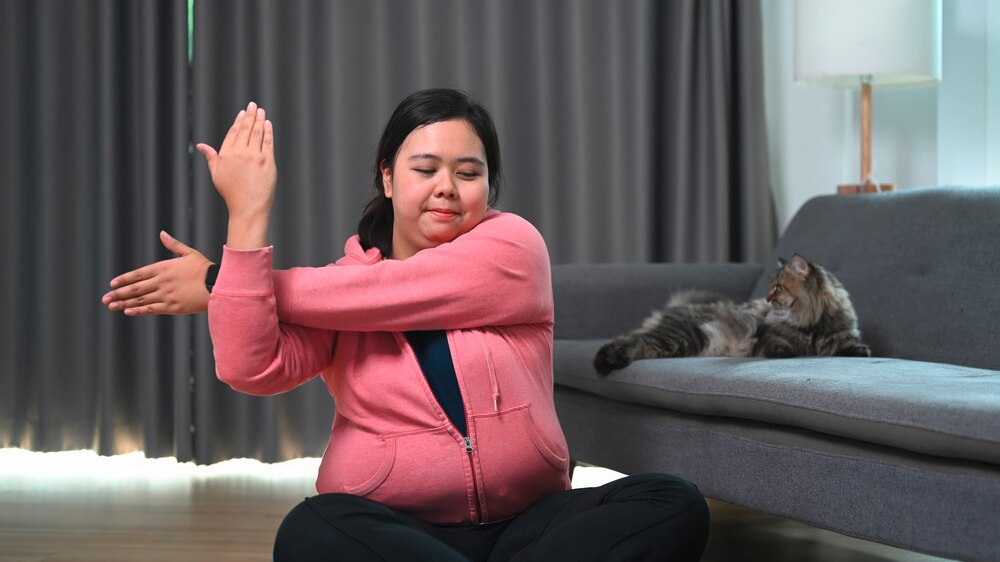 Asian woman in pink and black outfit doing stretches next to sofa with cat.