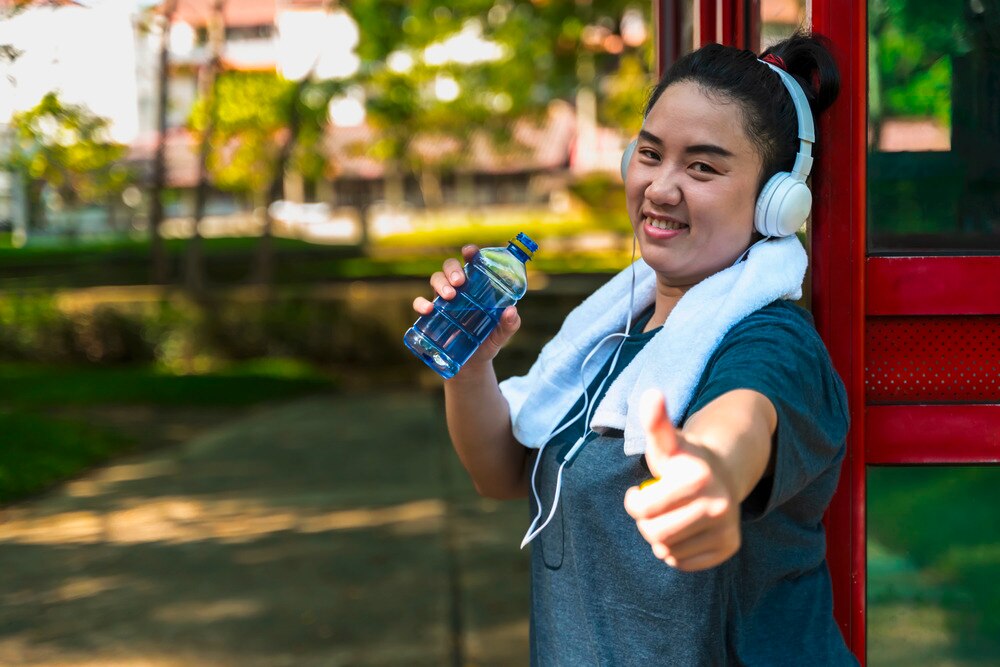 Asian woman with thumbs up sign, drinking water and wearing headphones.