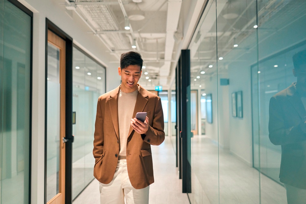 Man in brown blazer smiling while looking at his phone in the office.