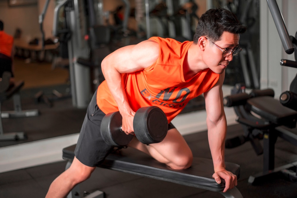Man in orange sleeveless top doing bent-over dumbbell row on a bench.